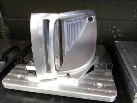 Molde metálico usinado com cavidade de formato curvo e aletas de refrigeração, fixado em mesa de máquina CNC